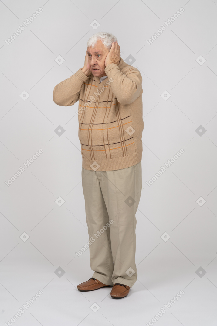 Front view of a scared old man in casual clothes covering ears with hands