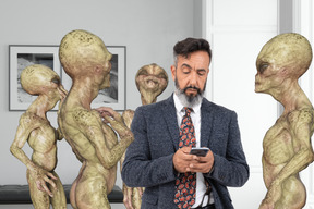 Man in suit and tie looking at cell phone while surrounded by group of aliens