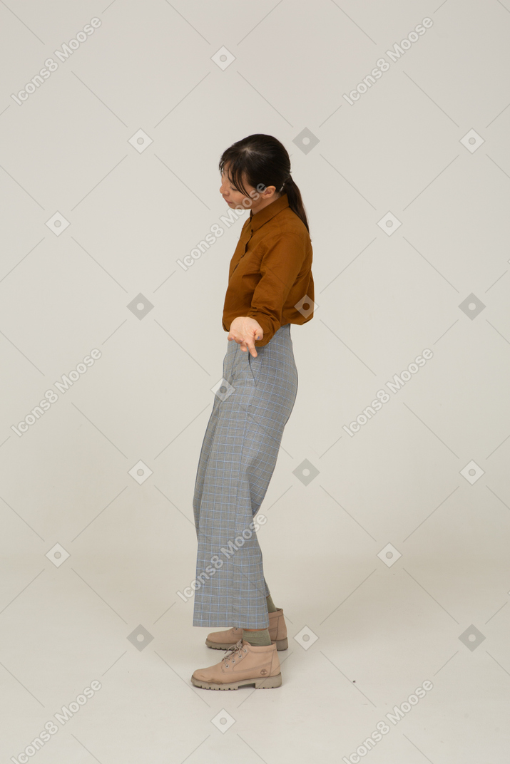 Vista lateral de uma jovem mulher asiática de calça e blusa, inclinando a cabeça e estendendo a mão