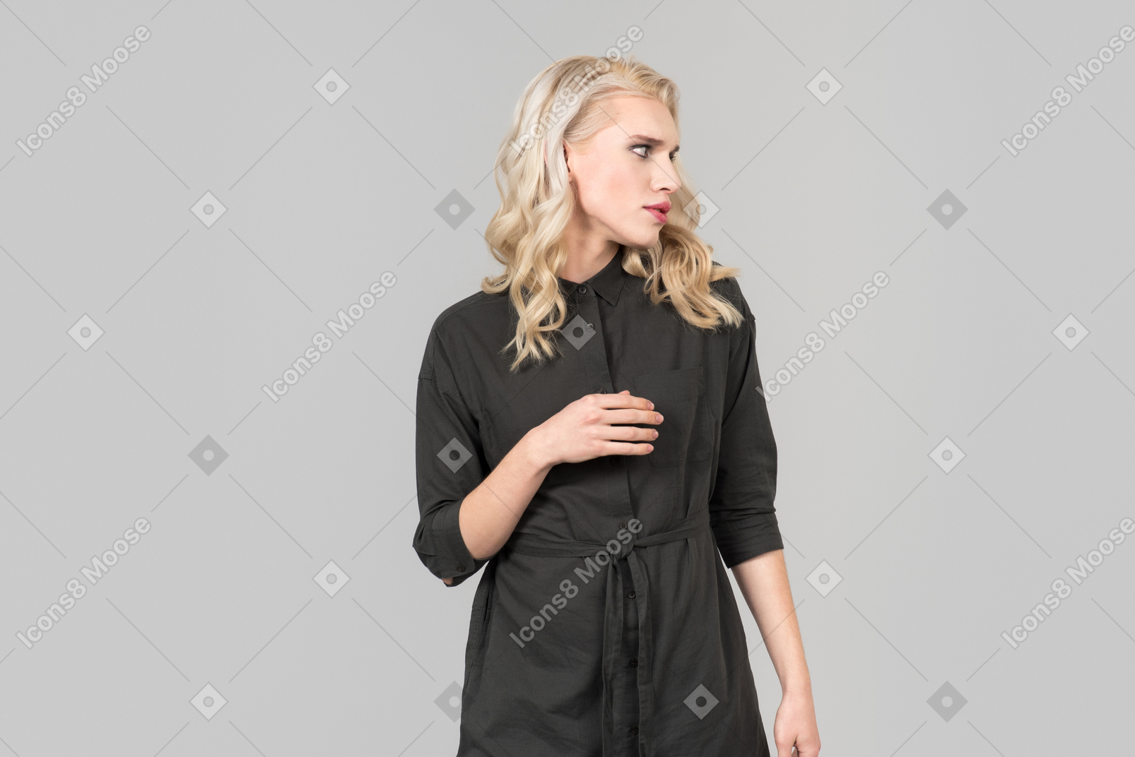 Una giovane persona dai capelli biondi in un abito nero in piedi contro il semplice sfondo grigio