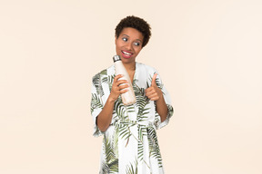 Young black short haired woman in a dressing gown, holding a reusable bottle and showing thumbs up