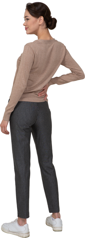 Vista posterior de tres cuartos de una mujer complacida en jersey y pantalones poniendo la mano en la cadera