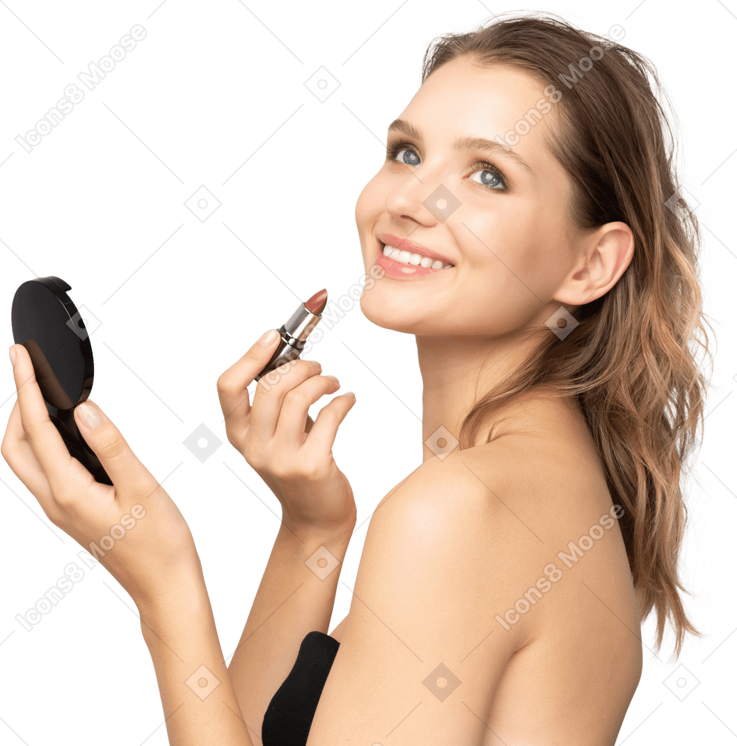 Vista lateral de una mujer joven sonriente aplicando lápiz labial mientras sostiene un espejo