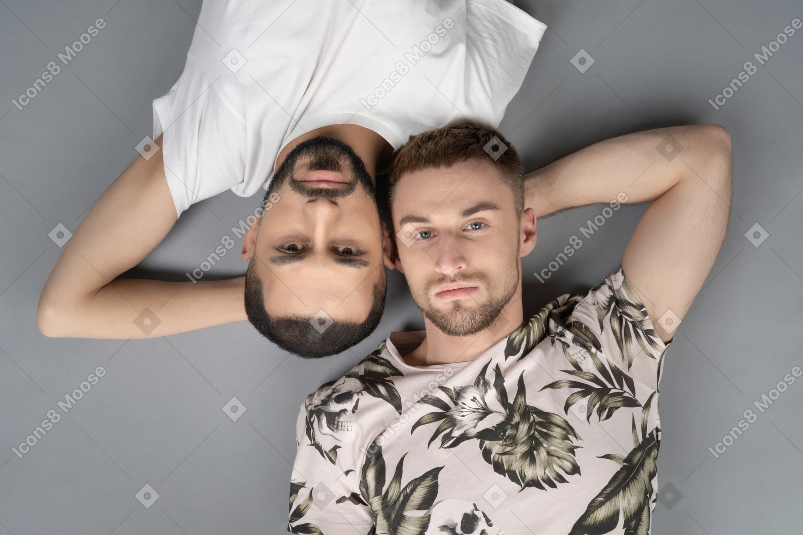 两个年轻的白人男子平躺着，头对头对称地躺着，看着相机
