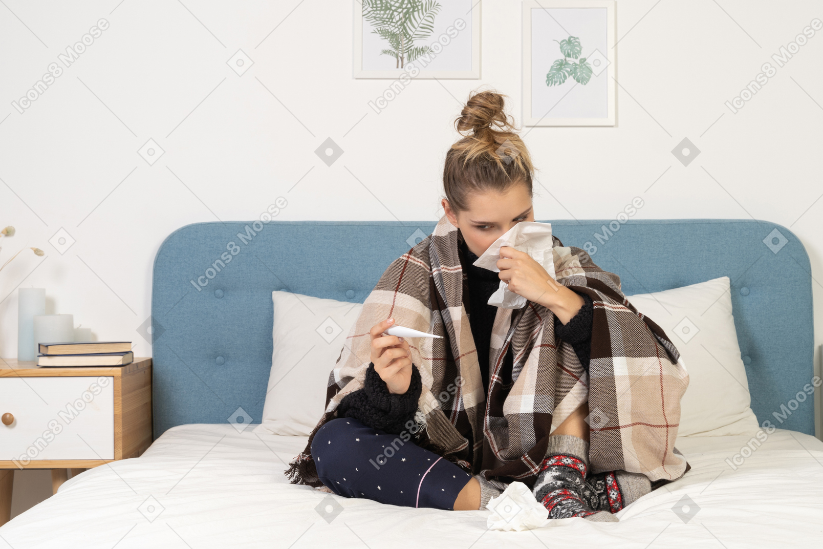 Front view of an ill young lady in pajamas wrapped in checked blanket blowing her nose