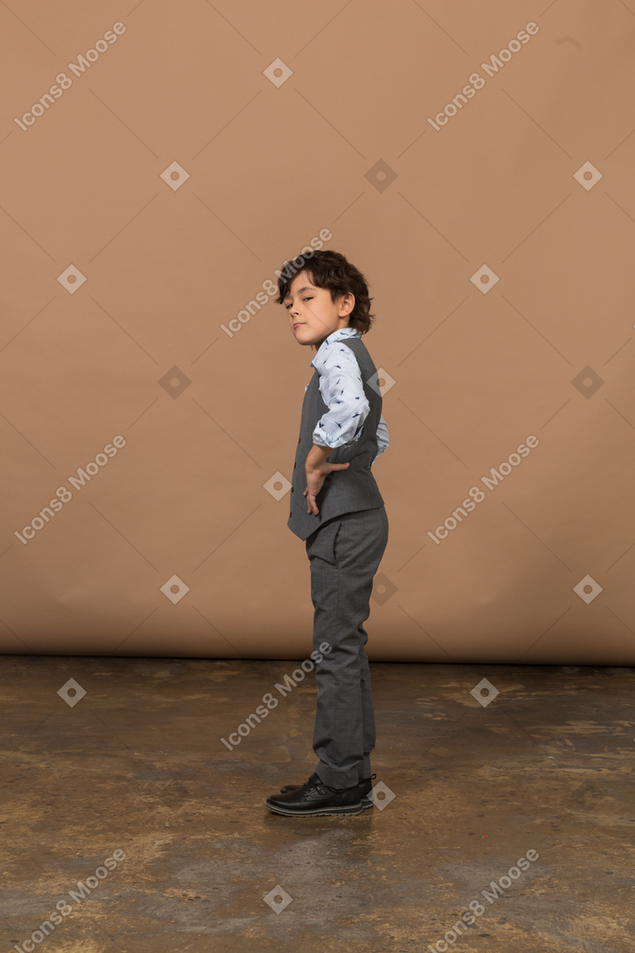 Side view of a cute boy in grey suit standing with hands on hips and looking up