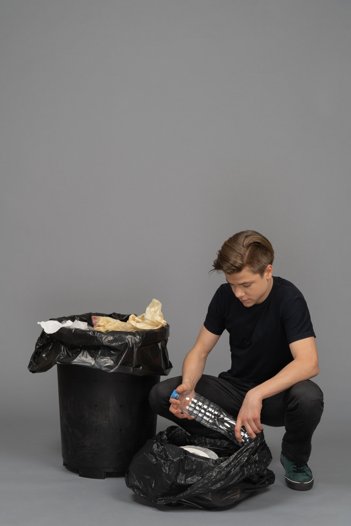 A young man sitting next to waste bin with plastic bottle in hands