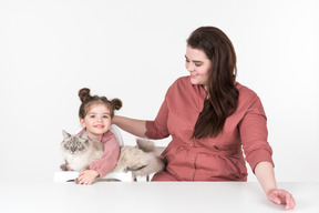 Mother and her little daughter, wearing red and pink clothes, sitting at the dinner table with their family cat