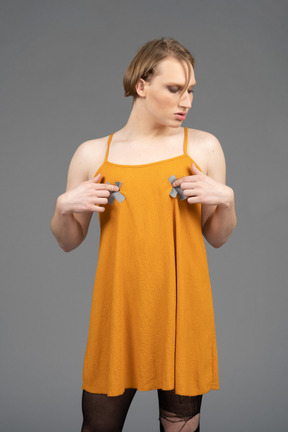 Молодой небинарный человек в оранжевом платье трогает грудь