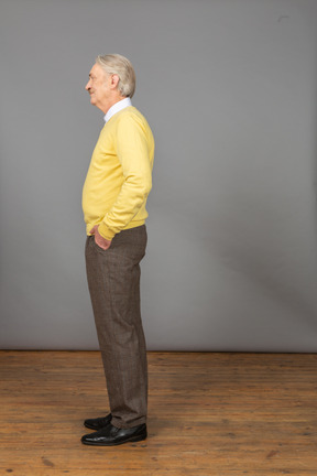 Side view of a displeased old man wearing yellow pullover and putting hand in pocket