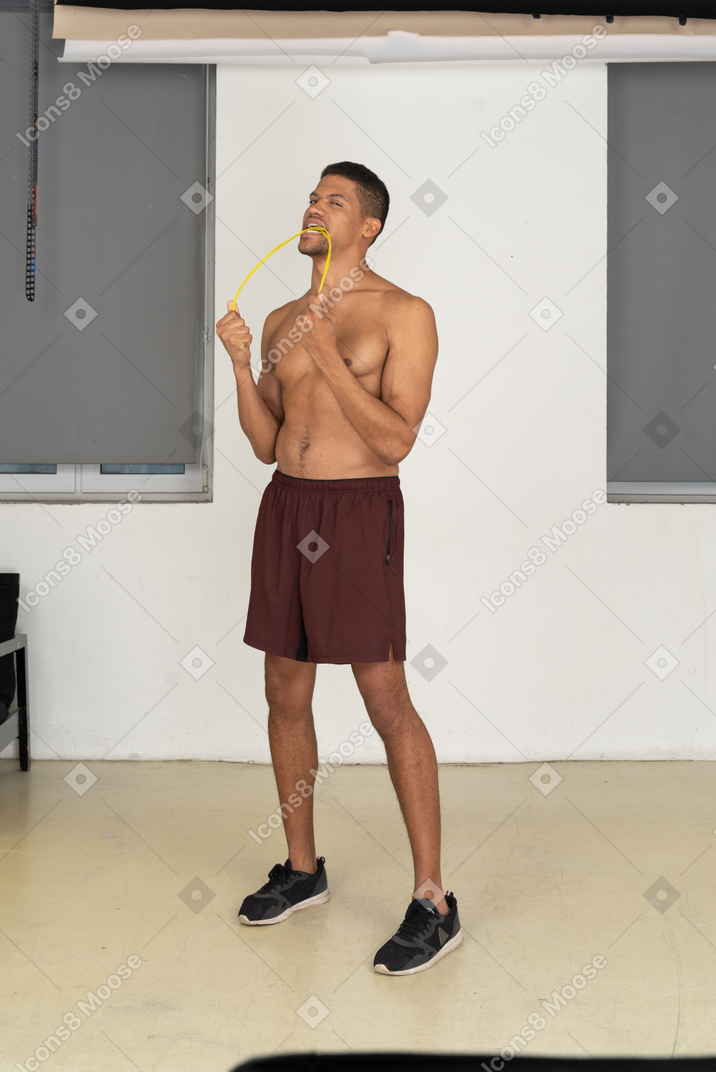 Young man in sport shorts standing half sideways and biting rope