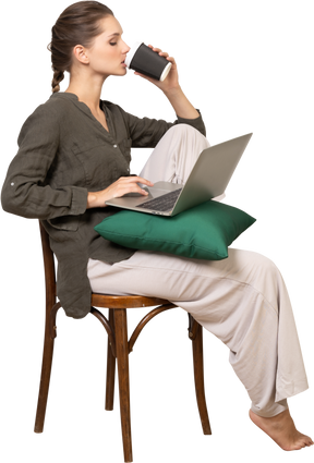 Вид сбоку озадаченной молодой женщины, сидящей на стуле и держащей свой ноутбук и пьющей кофе