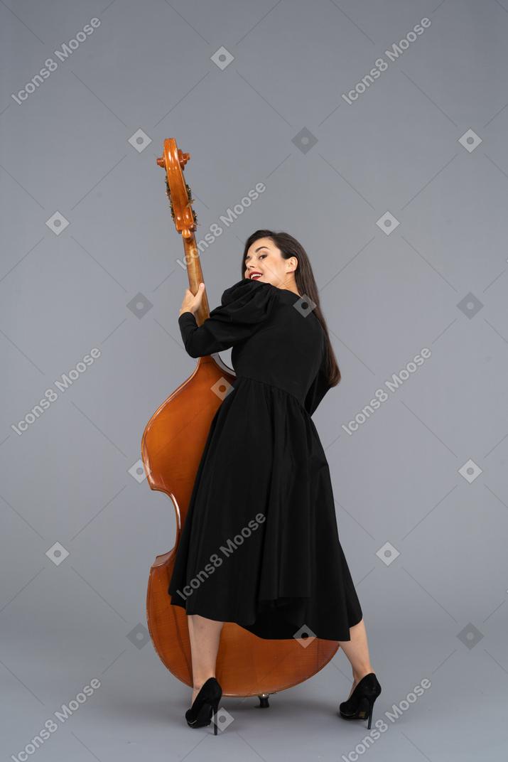 Vista posterior de un joven músico complacido en vestido negro sosteniendo su contrabajo
