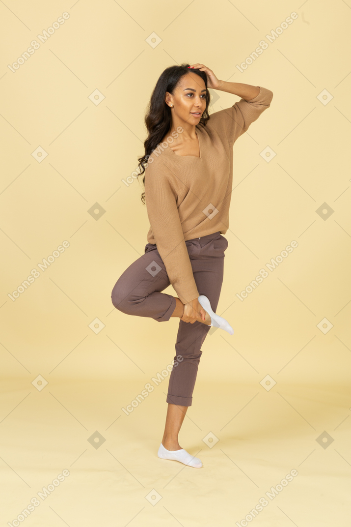 Three-quarter view of a dark-skinned young female raising leg while touching head