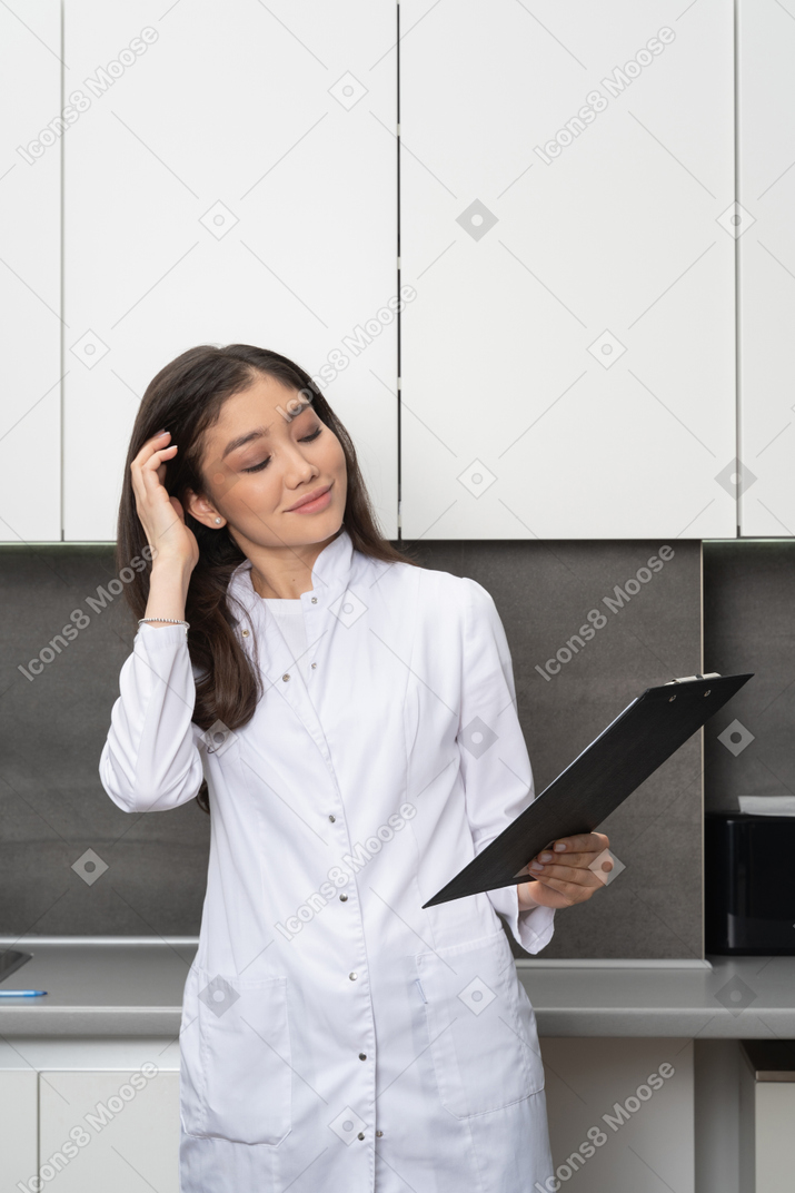 Front view of a pleased female doctor touching her hair and holding a tablet with her eyes closed