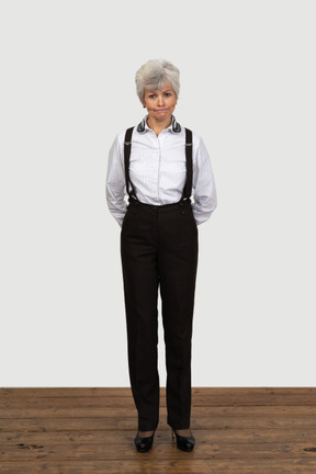 Vista frontal de una vieja mujer disgustada en ropa de oficina haciendo muecas con las manos detrás de la espalda