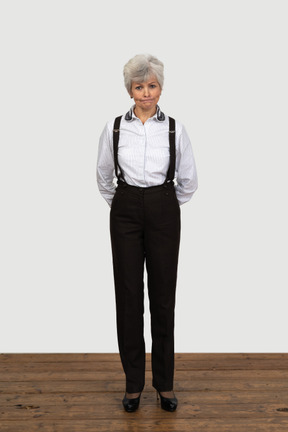 Front view of an old displeased female in office clothes grimacing with her hands behind back