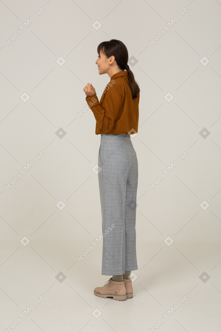 Vista posteriore di tre quarti di una giovane donna asiatica felice in calzoni e camicetta che alza le mani