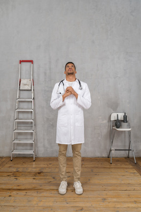 Front view of a praying young doctor standing in a room with ladder and chair