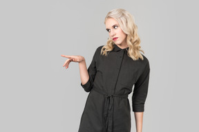 A young blond-haired person in a black dress  standing against the plain grey background