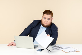 Молодой офисный работник с избыточным весом сидит за столом и держит документы