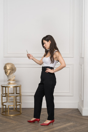 Vista lateral de uma jovem mulher olhando para o telefone e colocando a mão no quadril perto da escultura grega dourada