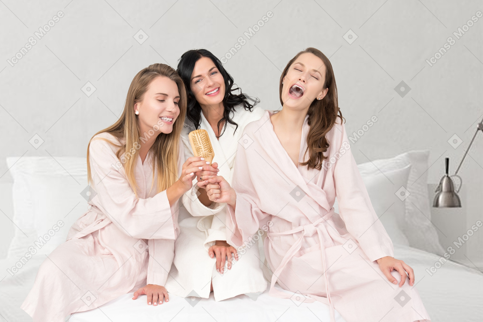 Three women in bathrobes sitting on a bed and singing with a hairbrush