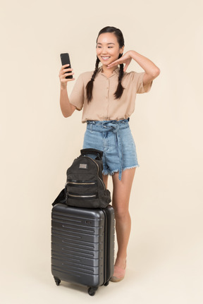 Smiling young woman standing near suitcase and making a selfie with a smartphone