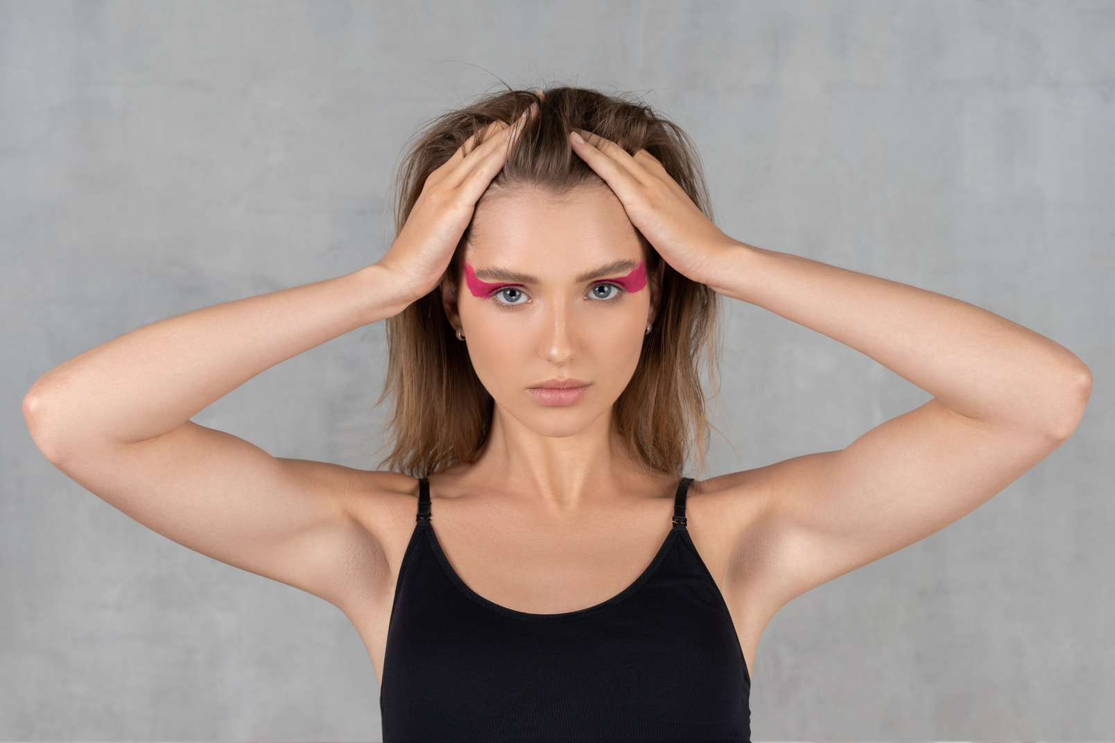 Front view of a young woman with bright pink eye make-up looking at camera & touching head