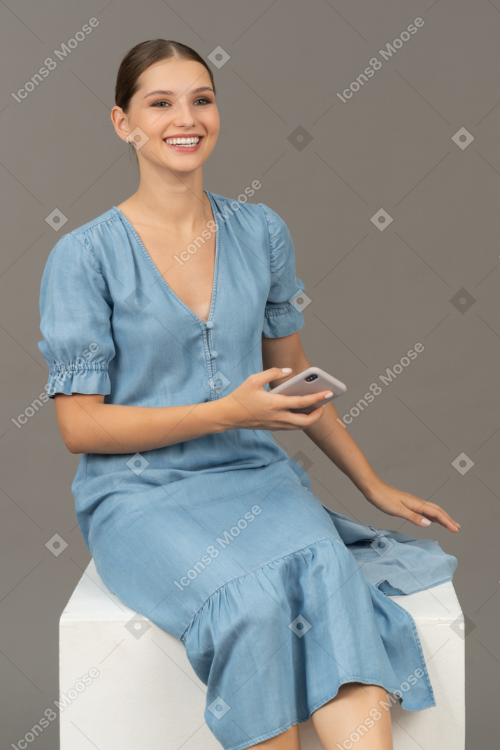 Three-quarter view of young woman sitting on a cube and smiling with smartphone in hand