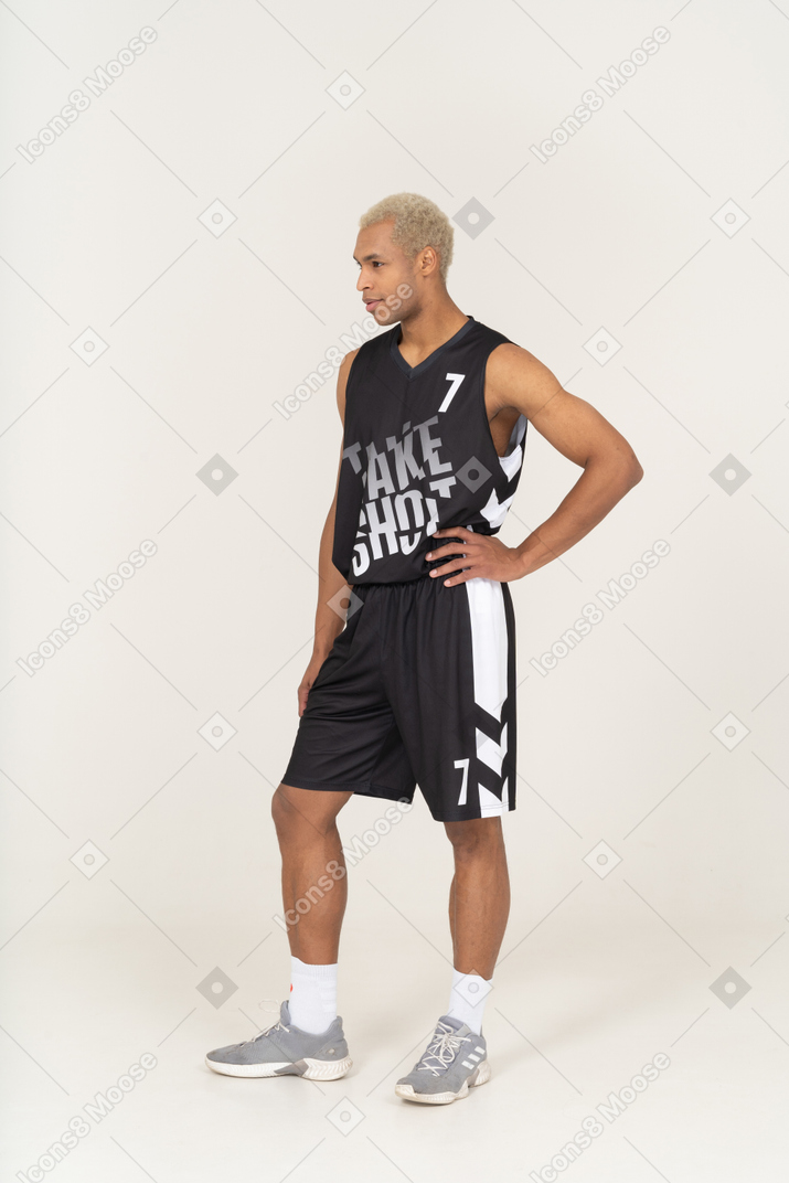 Three-quarter view of a young male basketball player putting hand on hip & looking aside