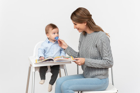 Young mom reading a book to her little son sitting in a highchair