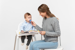 Jeune maman lisant un livre à son petit fils assis dans une chaise haute