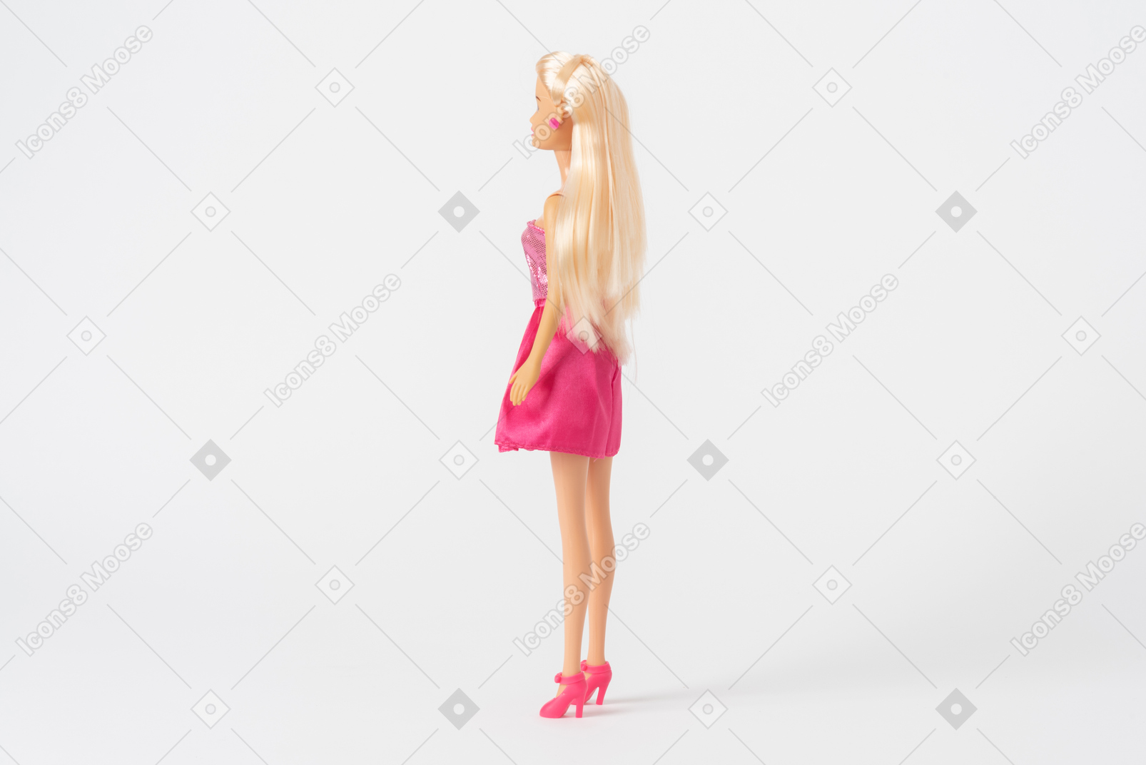 A side shot of a barbie doll in a shiny pink dress and pink high heels, standing isolated against a plain white background