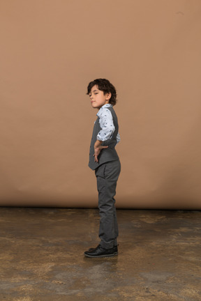 Vista lateral de un chico lindo con traje gris parado con las manos en las caderas y mirando hacia arriba