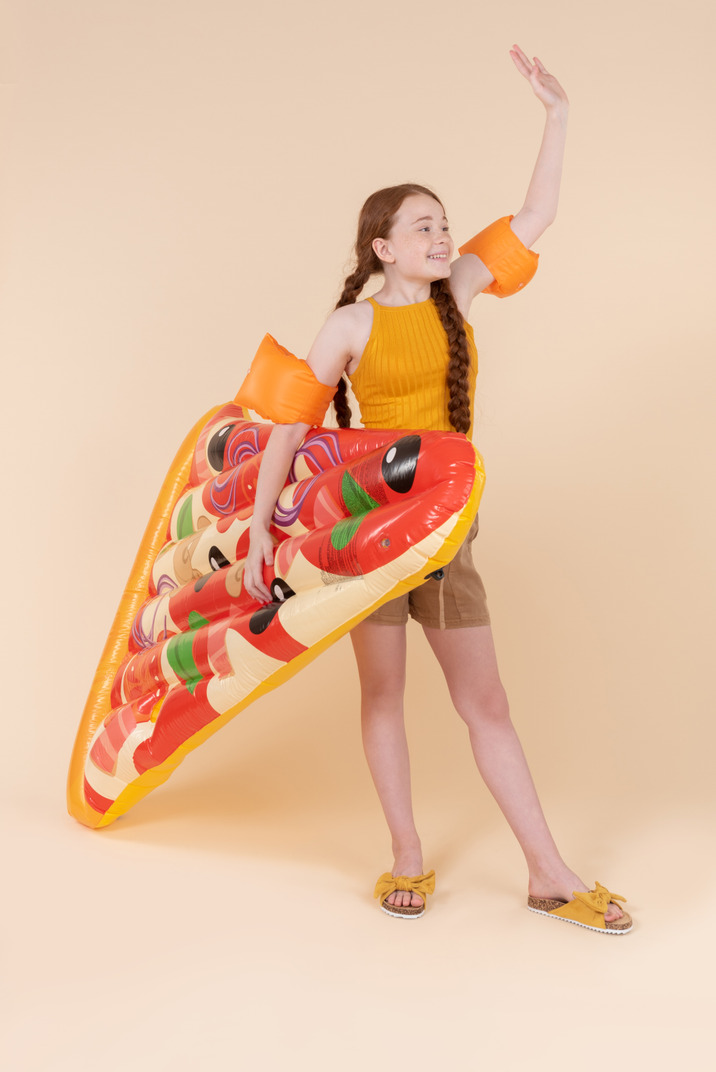 Teenage girl wearing arm floats, holding pizza mattress and waving