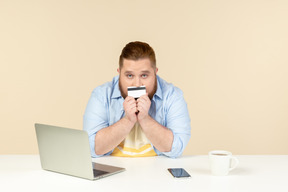 Young overweight man sitting at the table and holding bank card close to his mouth