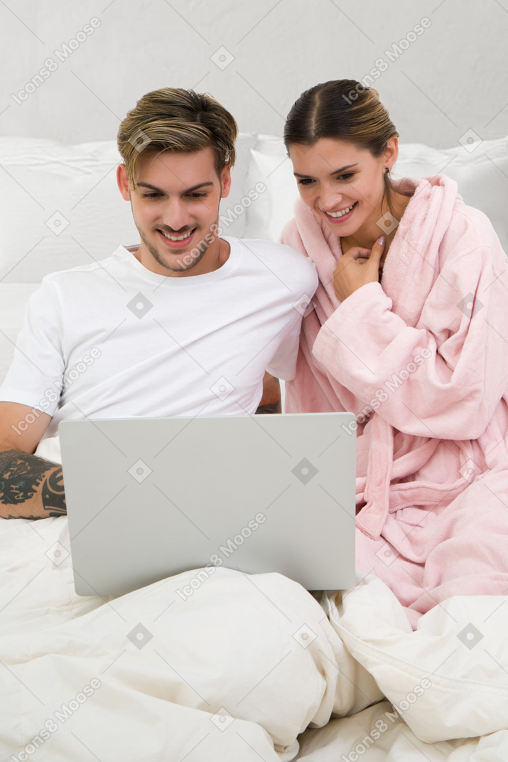 A man and woman sitting on a bed looking at a laptop