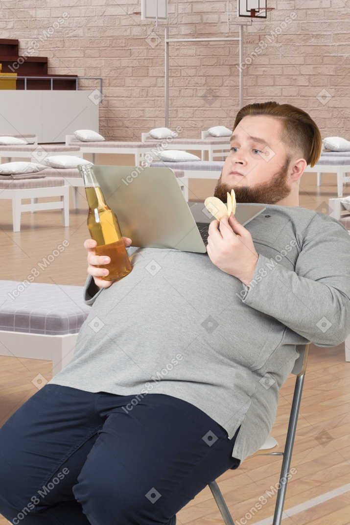 A man sitting on a chair with a bottle of beer and a laptop
