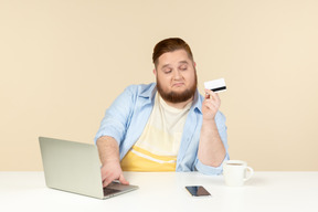 Серьезный молодой человек с избыточным весом сидит за домашним столом и держит банковскую карту