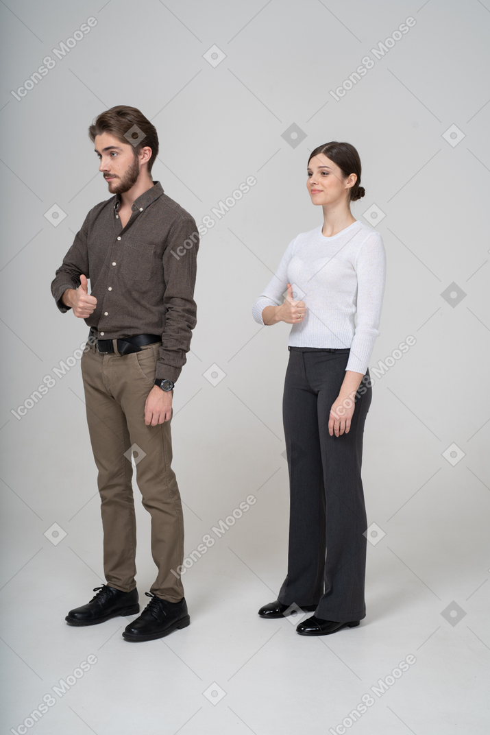 Vista de tres cuartos de una joven pareja alegre en ropa de oficina mostrando el pulgar hacia arriba