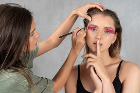 Vista frontal de una mujer joven que se maquilla mientras muestra un gesto de silencio
