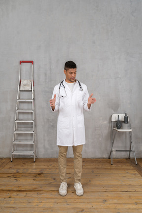 Front view of a young doctor standing in a room with ladder and chair showing a size of something