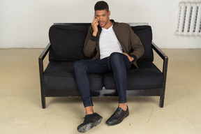 Front view of a perplexed young man sitting on a sofa and talking on his phone