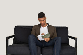 Vue de face du jeune homme assis sur un canapé et prenant des notes