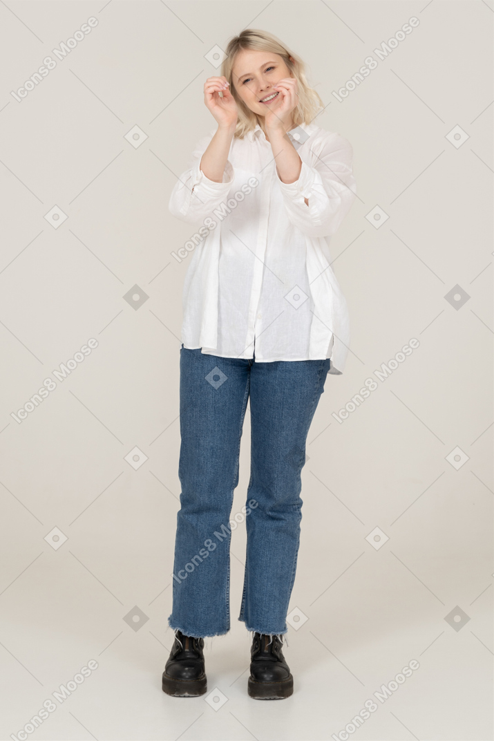 Front view of a blonde female in casual clothes showing a heart gesture and grimacing