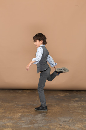 Vista lateral de un niño con traje gris posando en una pierna