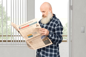 A bald man reading a newspaper while standing in front of a window