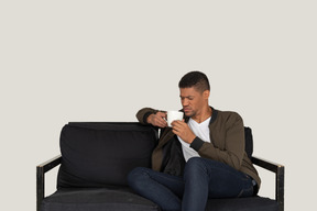 Front view of a young displeased man sitting on a sofa with a cup of coffee