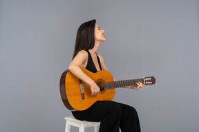 Вид сбоку сидящей улыбающейся молодой леди в черном костюме, играющей на гитаре